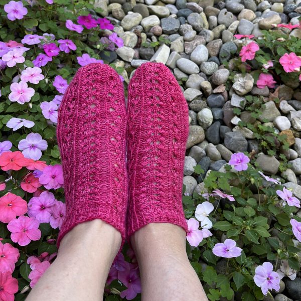 Two modelled short (trainer) socks with a twisted rib and lace pattern covering the top of the foot. The socks are knit in pink yarn.