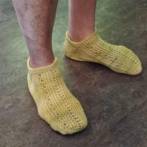 Two modelled short (trainer) socks with a twisted rib and lace pattern covering the top of the foot. The socks are knit in yellow yarn.