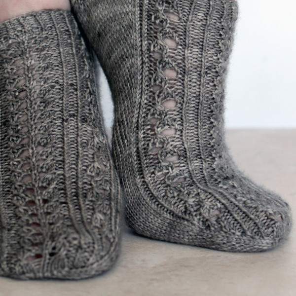 Two modelled short (trainer) socks with a twisted rib and lace pattern covering the top of the foot. The socks are knit in grey yarn.