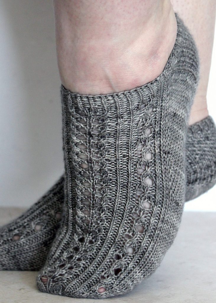 Two modelled short (trainer) socks with a twisted rib and lace pattern covering the top of the foot. The socks are knit in grey yarn.