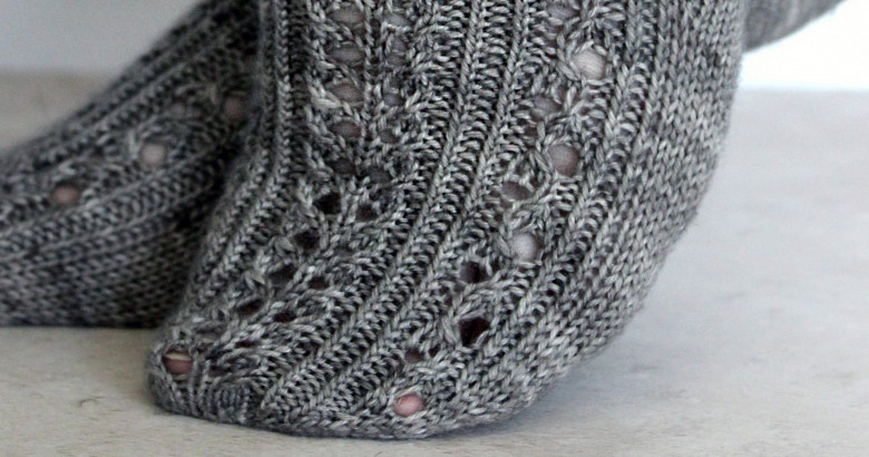 Two modelled short (trainer) socks with a twisted rib and lace pattern covering the top of the foot. The socks are knit in grey yarn.
