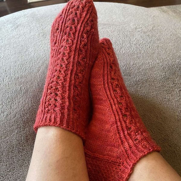 Two modelled short (trainer) socks with a twisted rib and lace pattern covering the top of the foot. The socks are knit in red yarn.